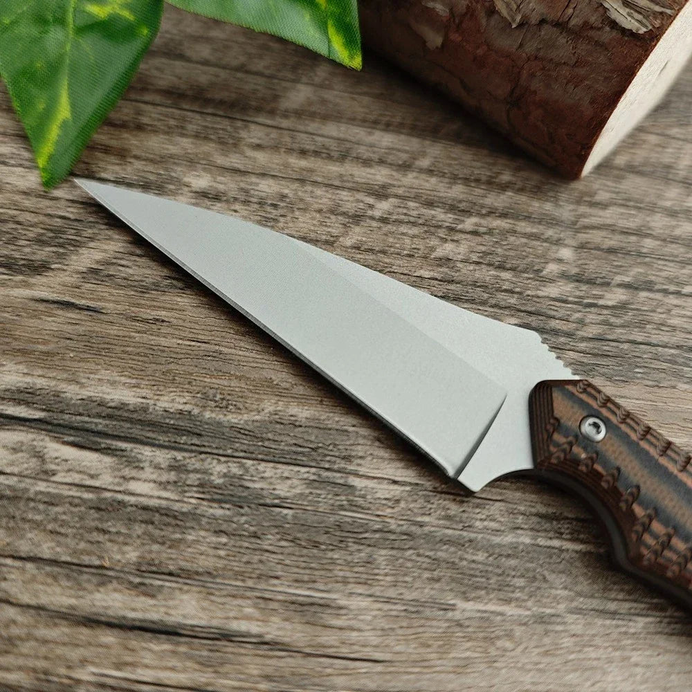 A silver compact fixed blade utility knife rests on a wooden surface next to a green leaf and a piece of wood.