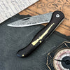 A folding Damascus steel knife with an ebony handle rests on aged parchment paper.