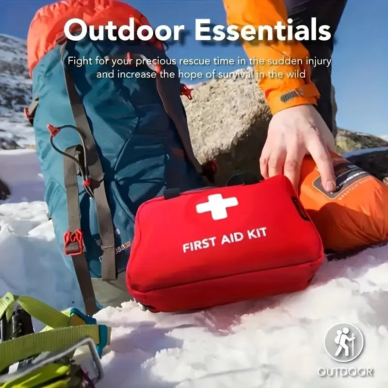 A red first aid kit is placed on snow next to an orange jacket and a green backpack in an outdoor setting.