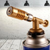 A brass professional gas torch with a metal body and nozzle is shown against a white brick background.