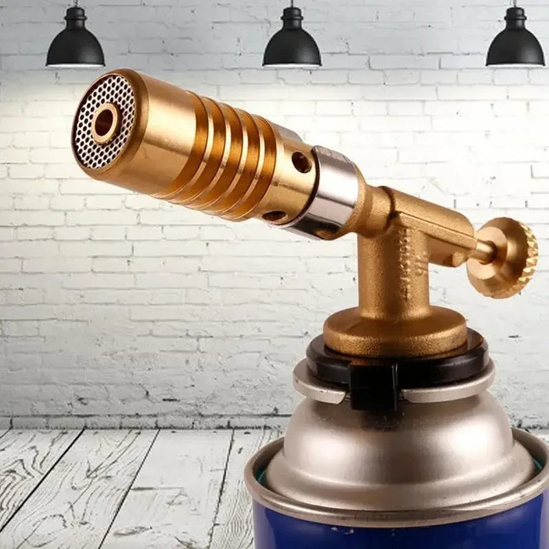 A brass professional gas torch with a metal body and nozzle is shown against a white brick background.
