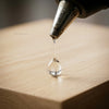 A drop of clear glue is dispensed from a hot melt glue gun onto a wooden surface.