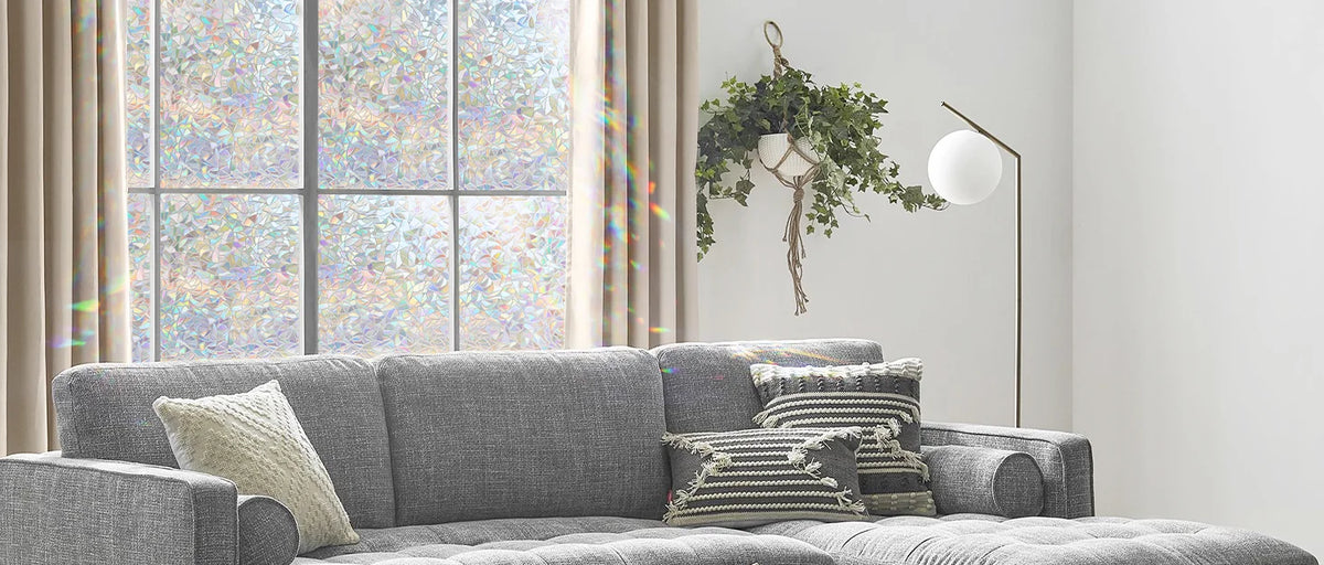 A gray sofa with patterned pillows sits in a bright room near a window decorated with green foliage and a white lamp.