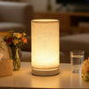 A retro style table lamp with a cylindrical fabric shade emits a warm, soft glow on a light-colored surface.