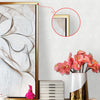 A framed abstract artwork with swirling lines in beige and white is displayed on a wall next to a gold-framed picture.