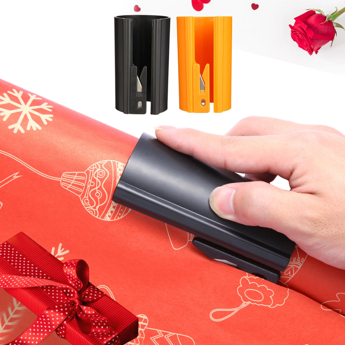 A hand holds a black sliding wrapping paper cutter next to orange and black versions, with red gift ribbon and rose visible.