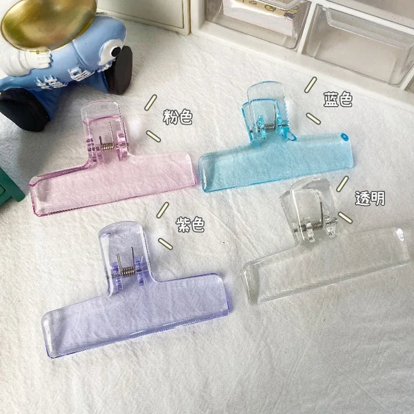 Various colored and clear transparent acrylic spring paper clips displayed on a white surface.