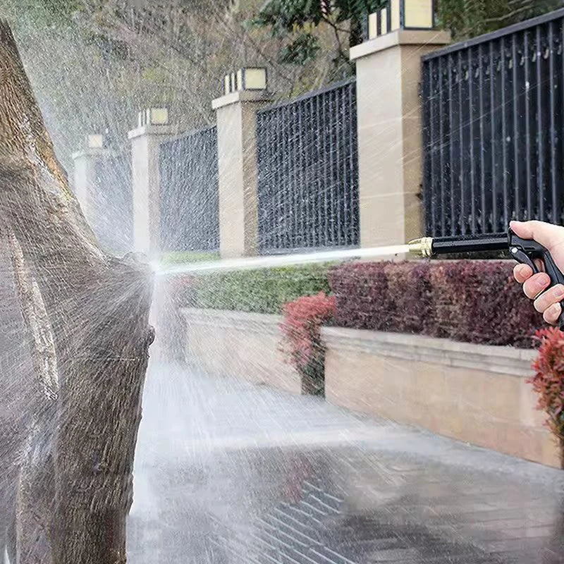 A hand sprays water from an expandable high-pressure garden hose onto a tree in a landscaped outdoor setting.