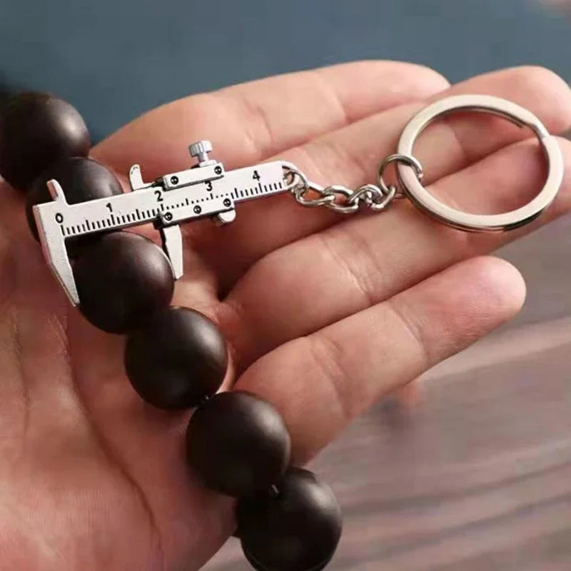 A hand holds a black beaded bracelet with a metal measuring ruler and caliper keychain attached.