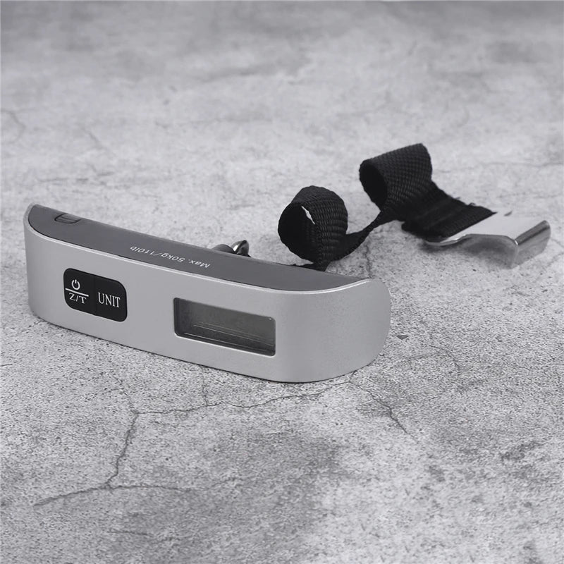 A silver portable digital weighing scale unit with a black strap is shown on a gray textured surface.