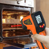 A hand holds an orange digital infrared thermometer gun measuring food inside an oven.