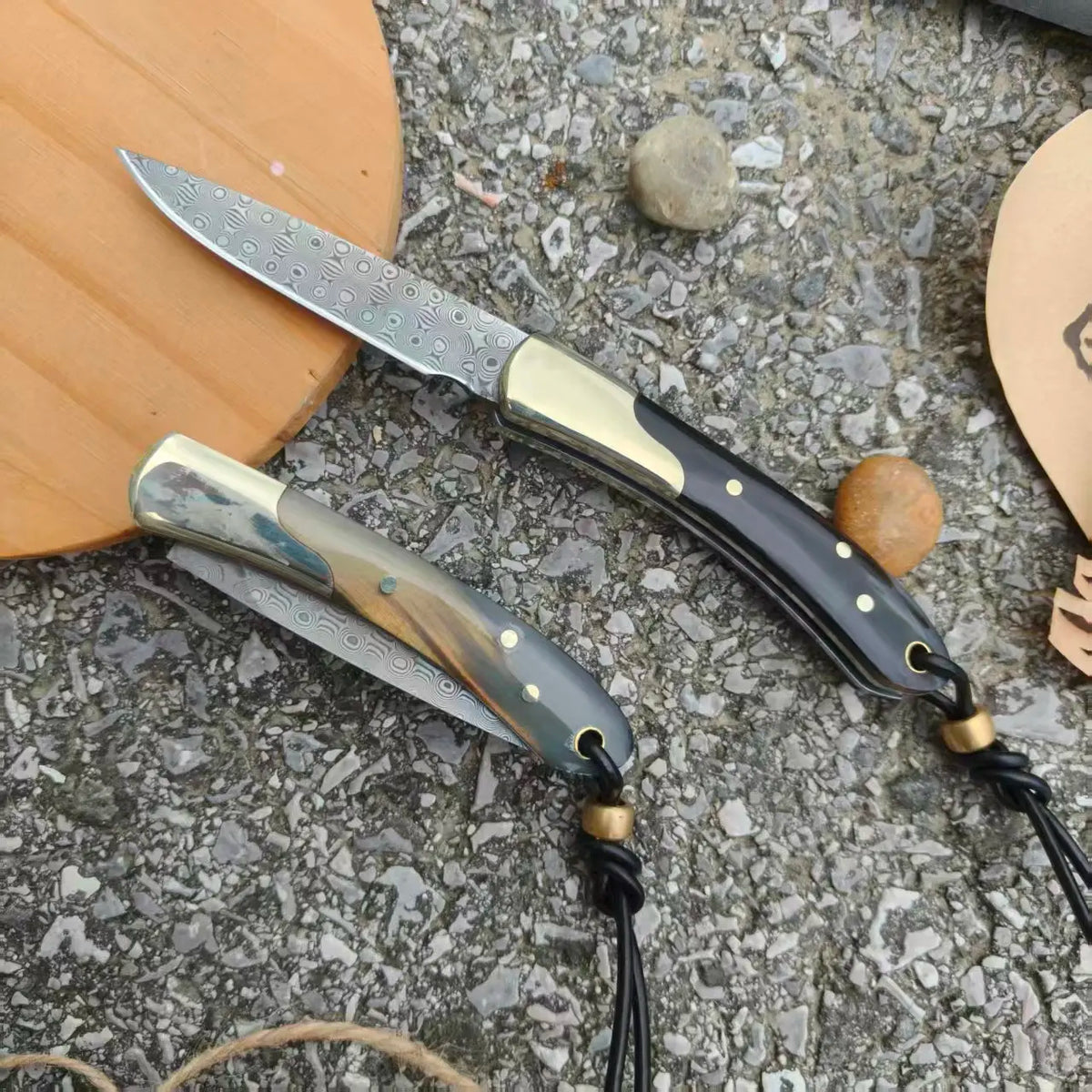 Un couteau en acier Damas pliable avec une lame argentée et un manche naturel foncé repose sur une surface de pierre.