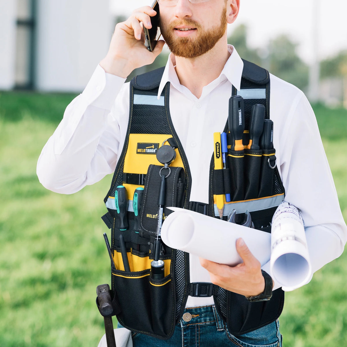 Un hombre con camisa blanca y chaleco de seguridad sosteniendo un teléfono al aire libre.