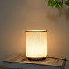 A minimalist desk lamp with a cylindrical fabric shade emitting a warm glow on a white surface.