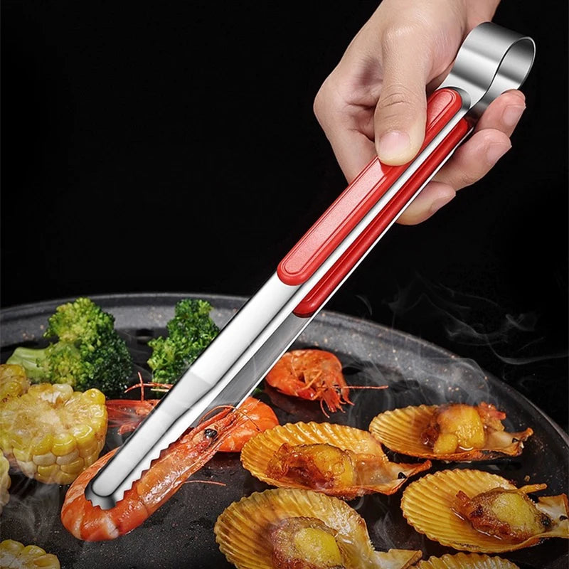 A hand holds a stainless steel BBQ grill tongs and kitchen clips set used for grilling seafood on a dark pan.