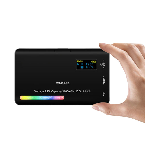 A hand holds a black RGB video light kit with display showing battery and power information.