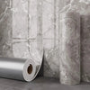 Rollo de papel autoadhesivo de mármol plateado exhibido contra una pared de mármol gris texturizada.