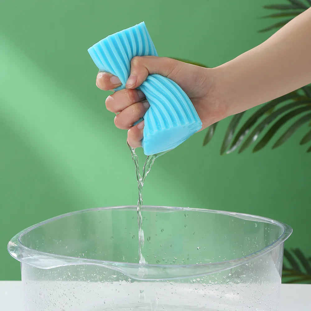 A hand is squeezing a blue PVA absorbent cleaning sponge into a clear bowl of water.