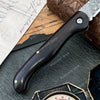 A close-up of a Damascus steel folding knife with an ebony handle resting on dark slate and parchment paper.
