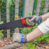 A hand wearing safety gloves is using a black blade to cut a wooden branch outdoors.