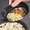 A hand is pressing a manual stainless steel garlic and ginger press, releasing minced garlic and ginger onto a dark plate.