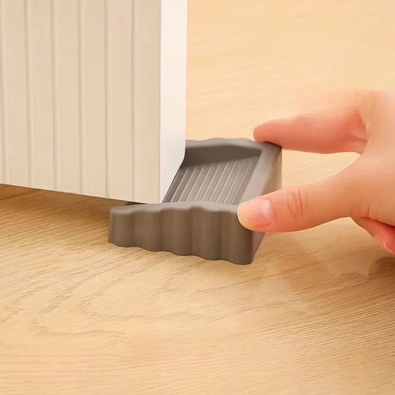A hand is holding a gray reusable TPR door stop wedge against a white door frame.