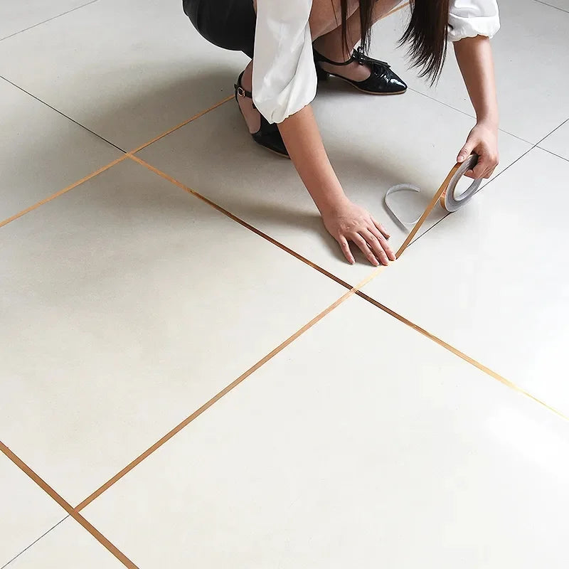 A person's hand is applying gold self-adhesive decorative tile gap tape to a white floor surface.
