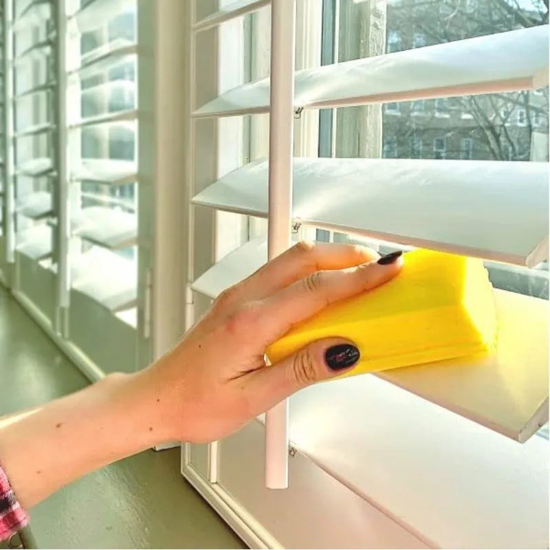 A hand is placing a yellow PVA absorbent cleaning sponge onto a white shelf inside a window frame.