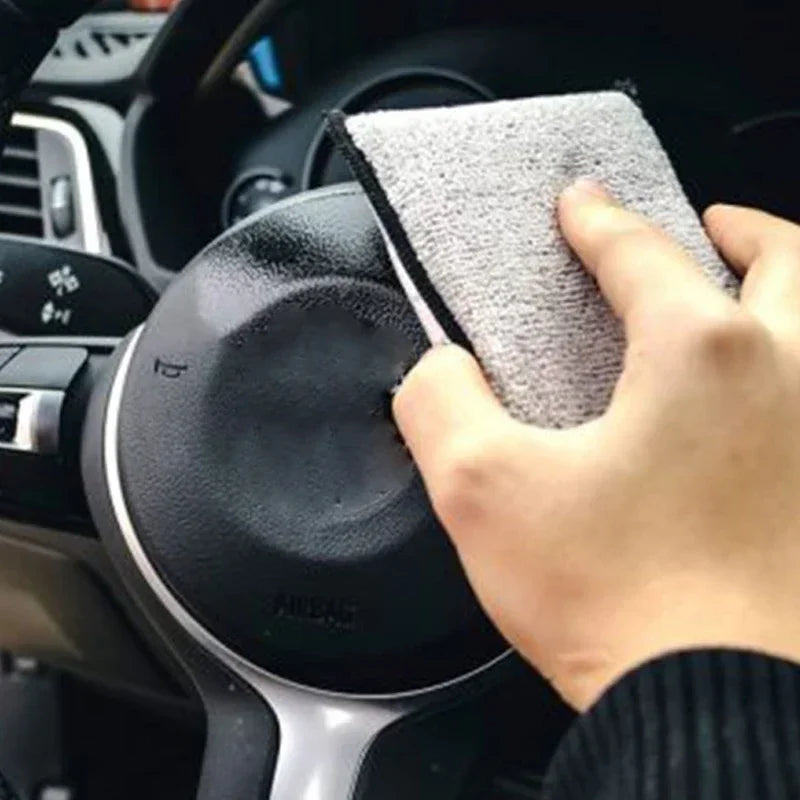 A hand is holding a gray microfiber detailing sponge against a black car interior surface.