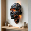 A dark, cracked face sculpture made of resin with glowing orange lines mounted on a white wall.