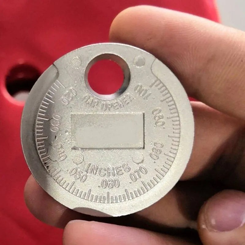 A hand holds a silver precision spark plug gap gauge with markings for measuring in inches.