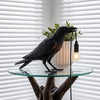 Eine schwarze Vogelstatue sitzt auf einer Glastischlampe mit Glühbirne vor einer Fensterblind.