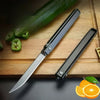 A black outdoor folding knife is displayed on a wooden board next to sliced green chili peppers and an orange.