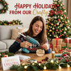 A woman is crafting holiday decorations with a cordless hot glue gun and various greenery on a wooden table.