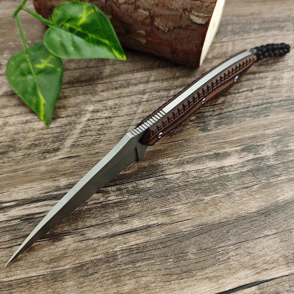 A compact fixed blade utility knife with a dark handle and silver blade resting on a wooden surface next to a green leaf.