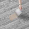 A hand is placing a square peel and stick wood grain floor tile onto a gray wooden floor.