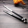 A pair of stainless steel BBQ grill tongs and kitchen clips resting on a dark wooden surface with a piece of fried food in th
