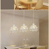 A modern crystal chandelier lamp with a white shade hangs above a wooden dining table in a bright room.