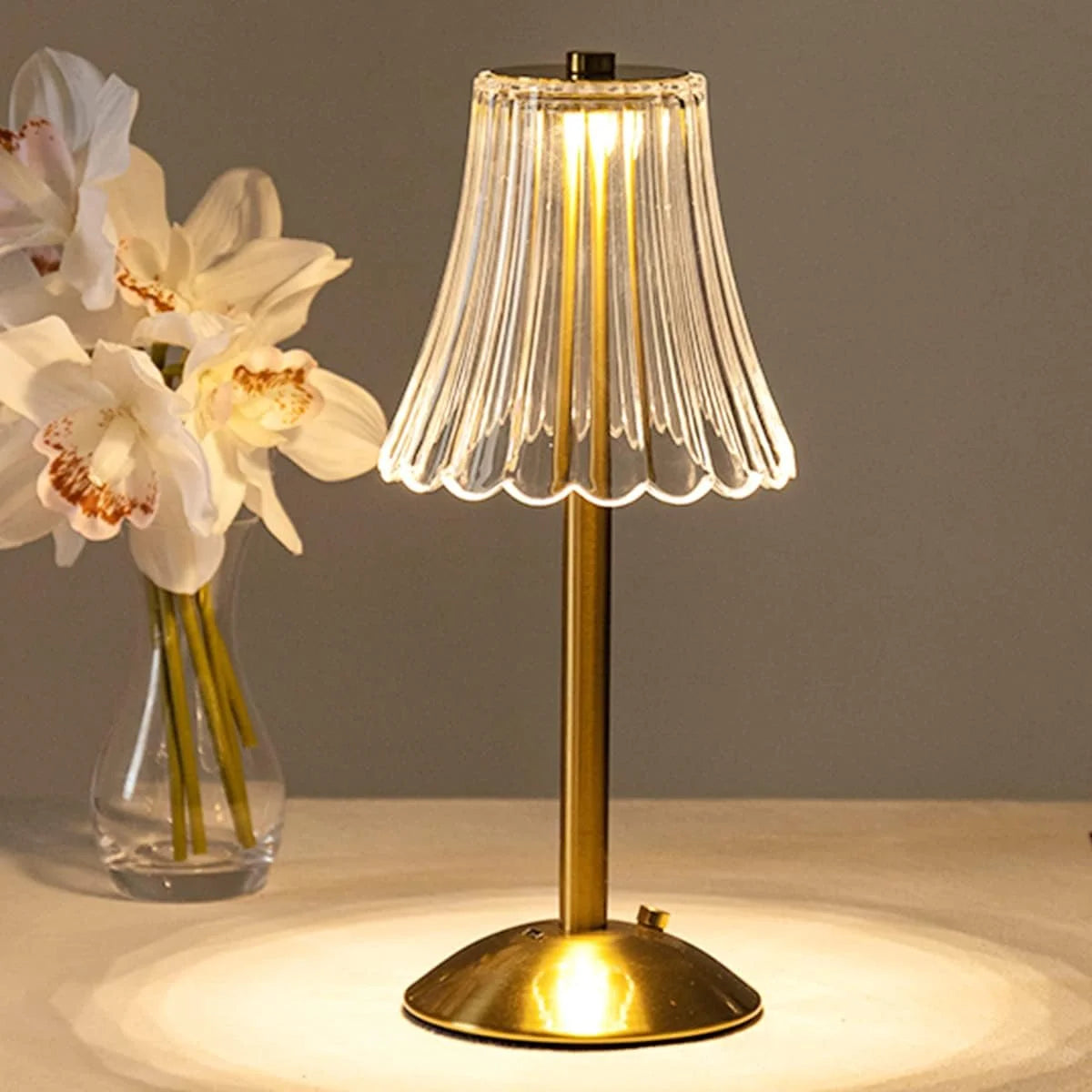 A brass table lamp with a clear pleated shade illuminates a white orchid flower.
