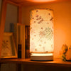 A retro style table lamp with a sheer fabric shade featuring nature-themed patterns on a wooden shelf.