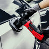 A hand wearing a glove is using a red compound buffing polishing pad on a car's exterior.