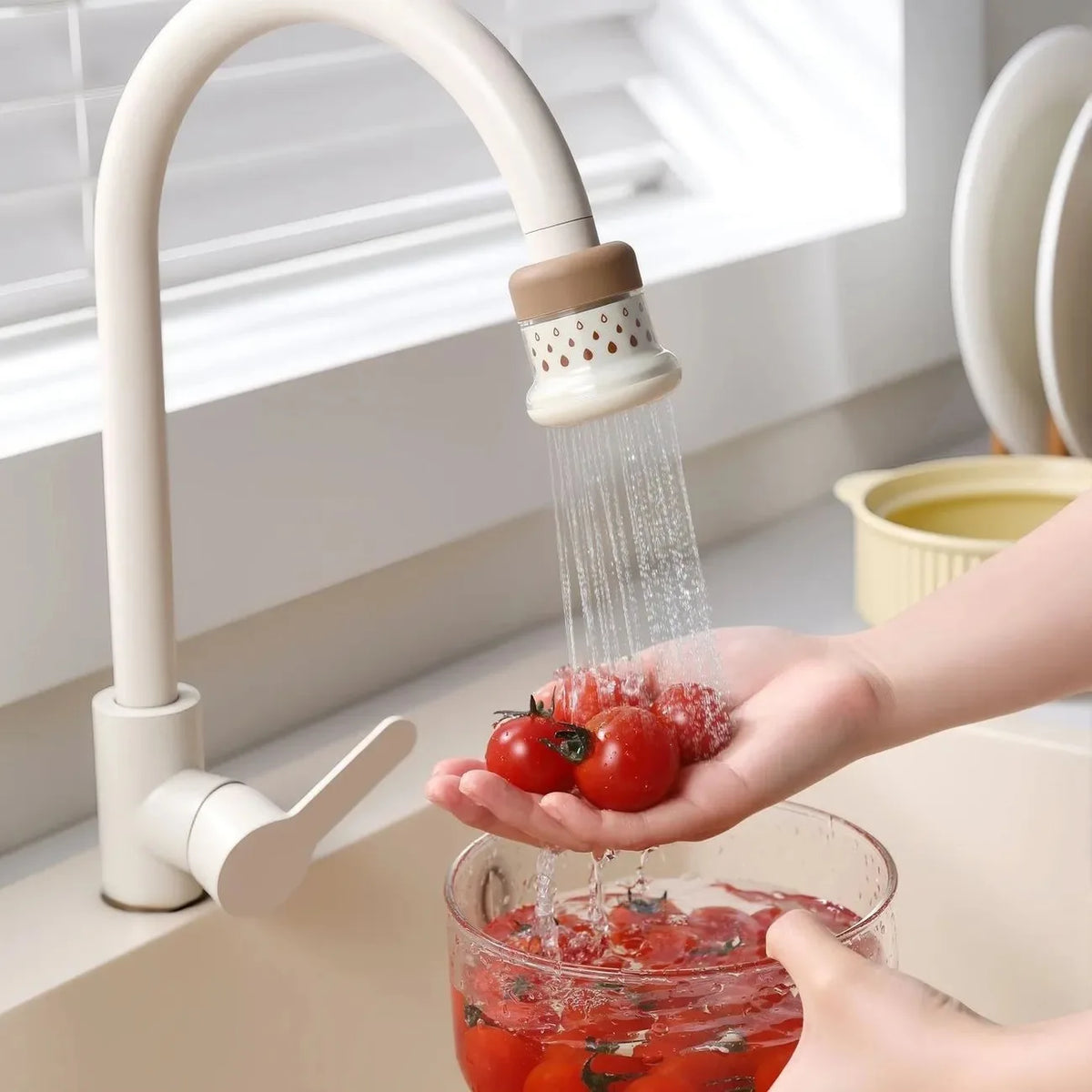 A hand is using an extendable tap filter sprayer to dispense water over fresh tomatoes in a glass.