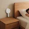 A wooden nightstand next to a bed with a white rotating electrical outlet extender mounted on the wall.