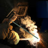 A hand holds a green welding rod, creating bright sparks and smoke in a dark environment.