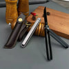 A black outdoor folding knife with a leather sheath and a silver blade is displayed on a wooden surface.