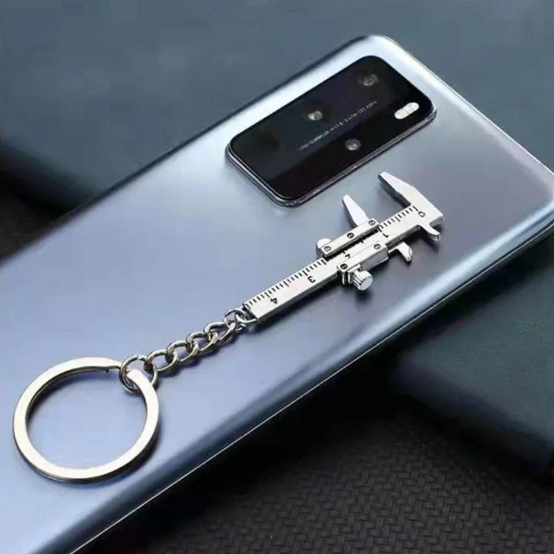 A silver metal jewelry measuring ruler and caliper keychain attached to a chain, placed on a smartphone.