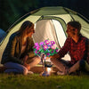 A couple sits in front of a tent with a glass lantern holding bright pink and purple solar LED azalea flowers.