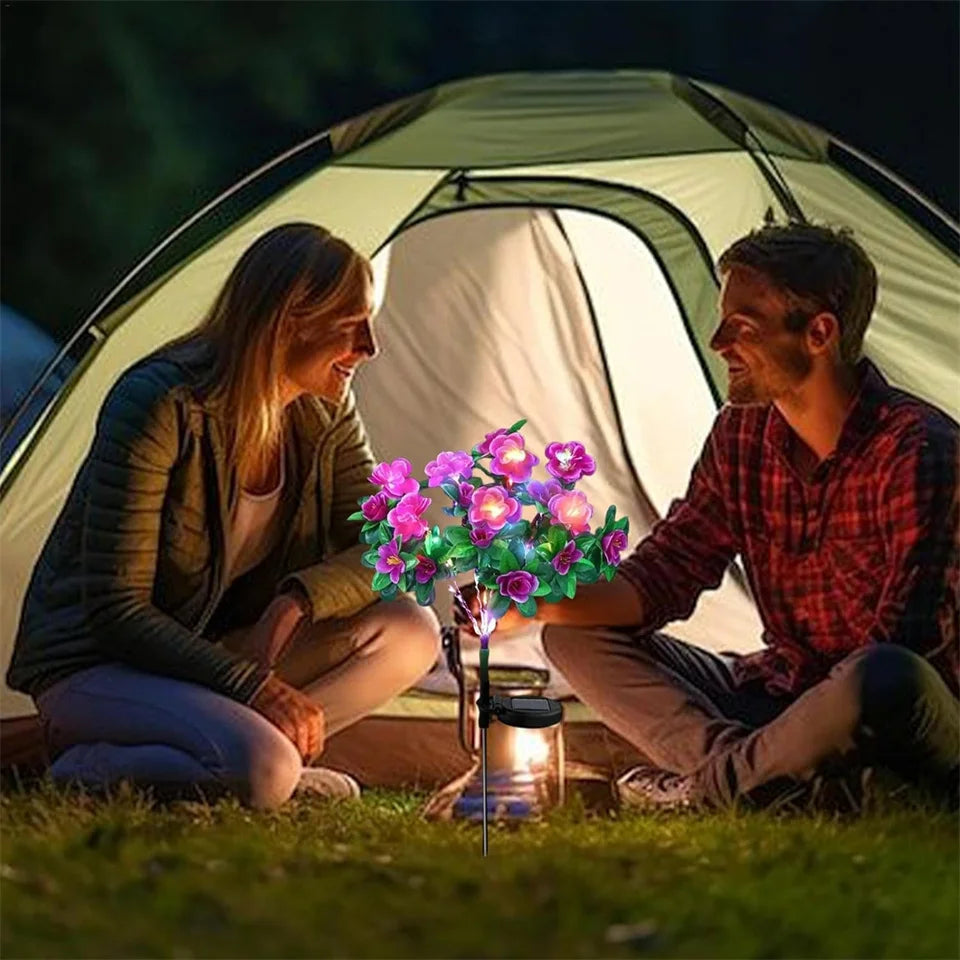 A couple sits in front of a tent with a glass lantern holding bright pink and purple solar LED azalea flowers.