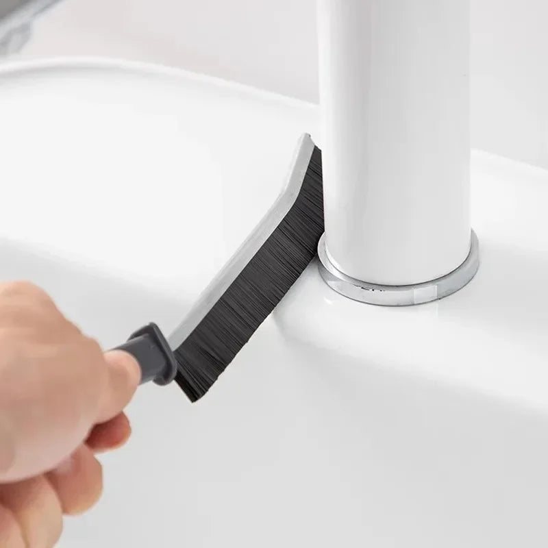 A hand is using a black grout gap cleaning brush to clean the base of a white cylindrical object.