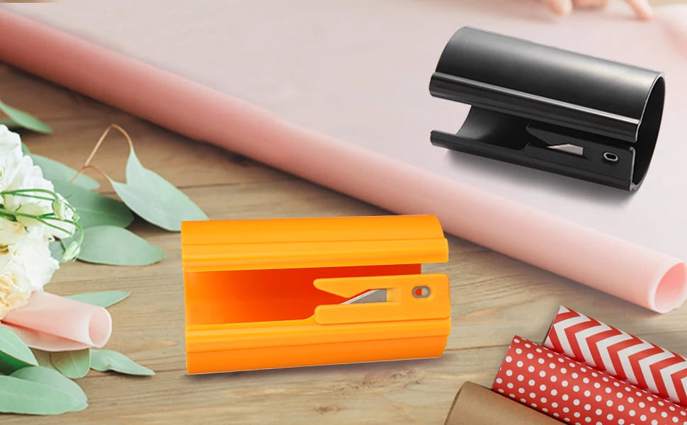 Orange sliding wrapping paper cutter and black cutter shown on a wooden surface with floral greenery.